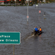 two people sit on a raft boat on a flooded street in front of a sign that reads "LaPlace" and "New Orleans"
